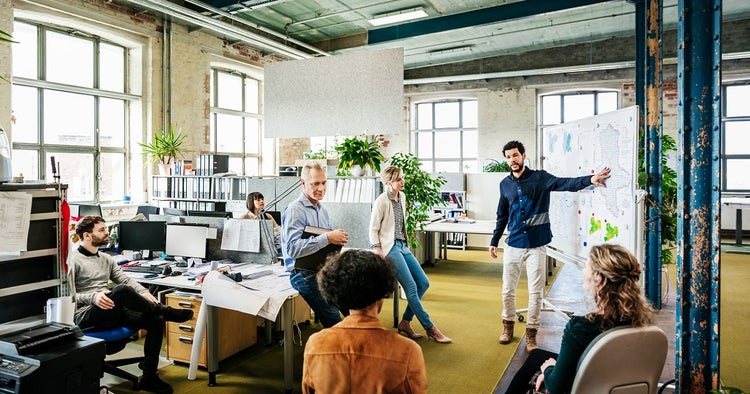 A manager is addressing his team during a morning meeting in a modern, open plan office environment.